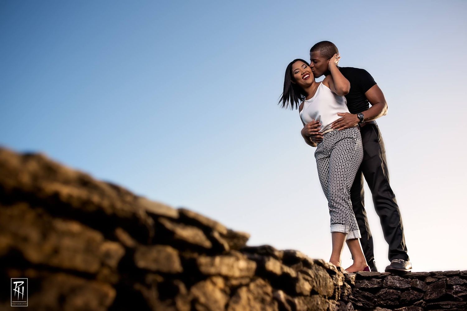 Couple sharing romantic moment on stone wall against blue sky background.