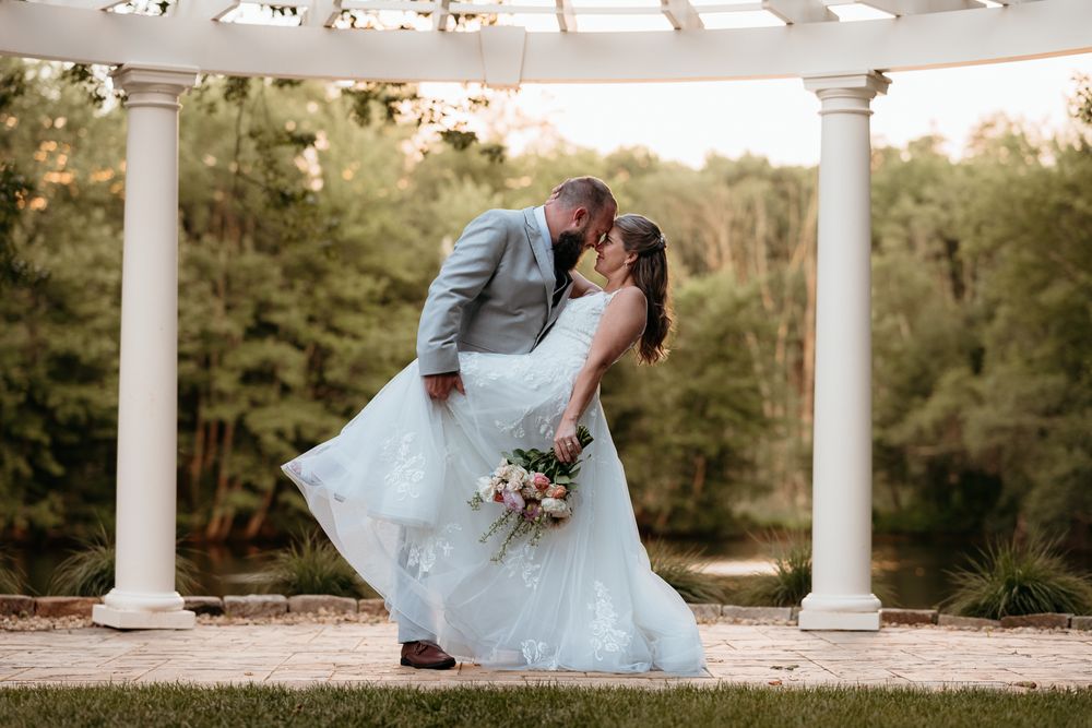 Bride and groom share a romantic dip kiss under a white pergola, surrounded by lush greenery at an outdoor wedding.