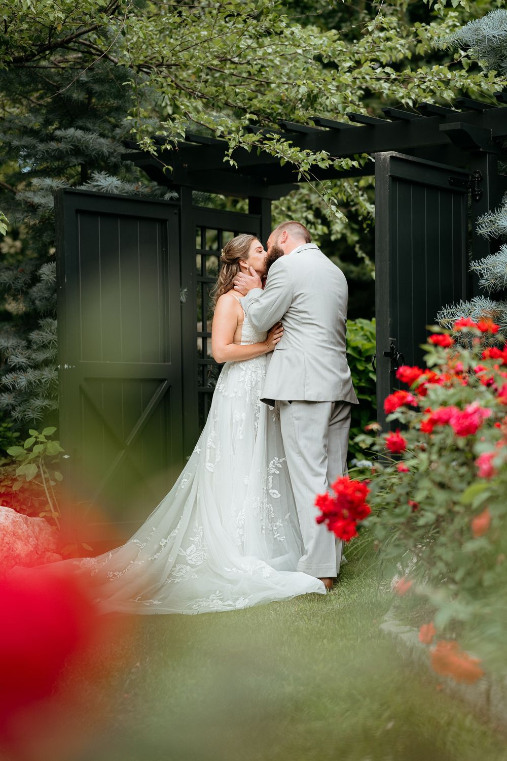 Bride and groom share a romantic kiss in a lush garden surrounded by red roses and a dark wooden gate.