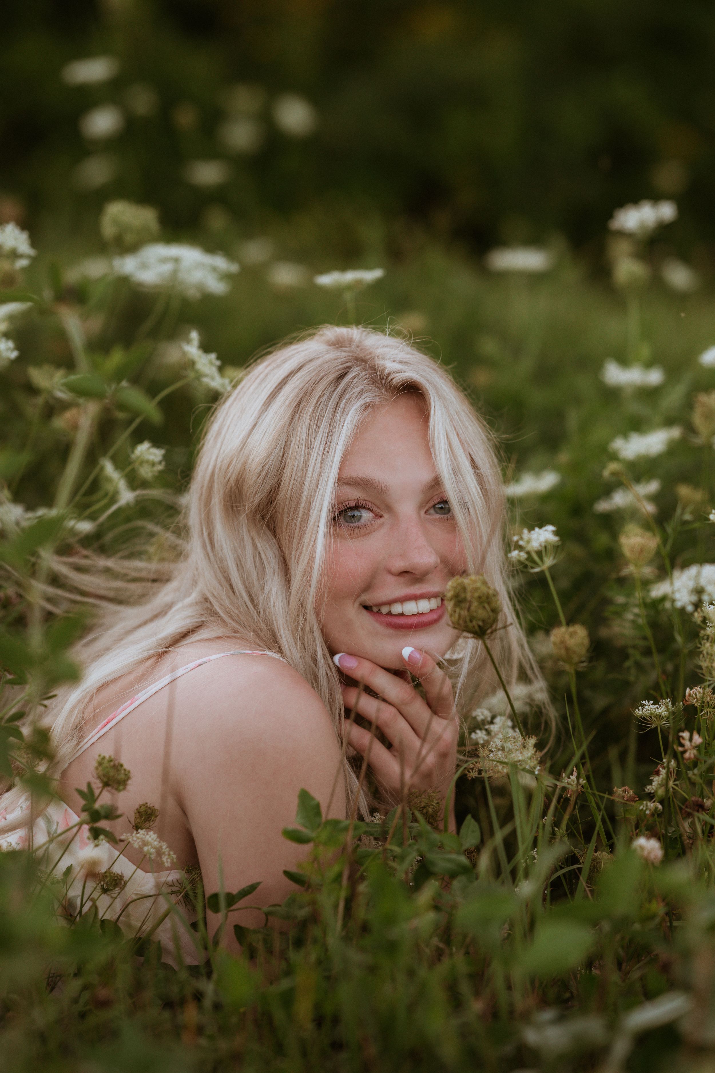 Smiling blonde woman lying in a wildflower field, holding a flower near her face with pink nails.