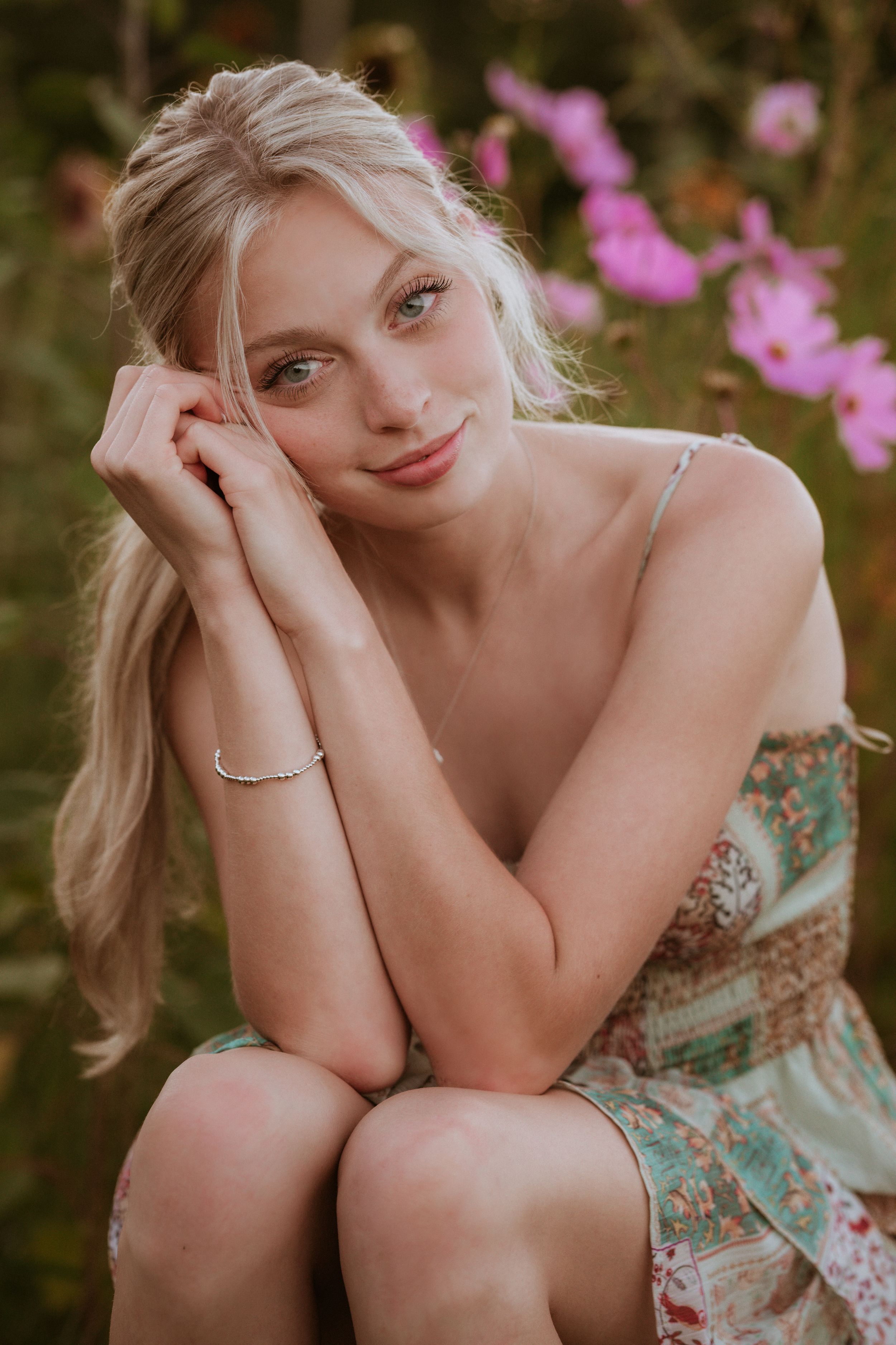 Blonde woman in floral dress sitting among pink cosmos flowers, smiling softly with hands clasped near face.