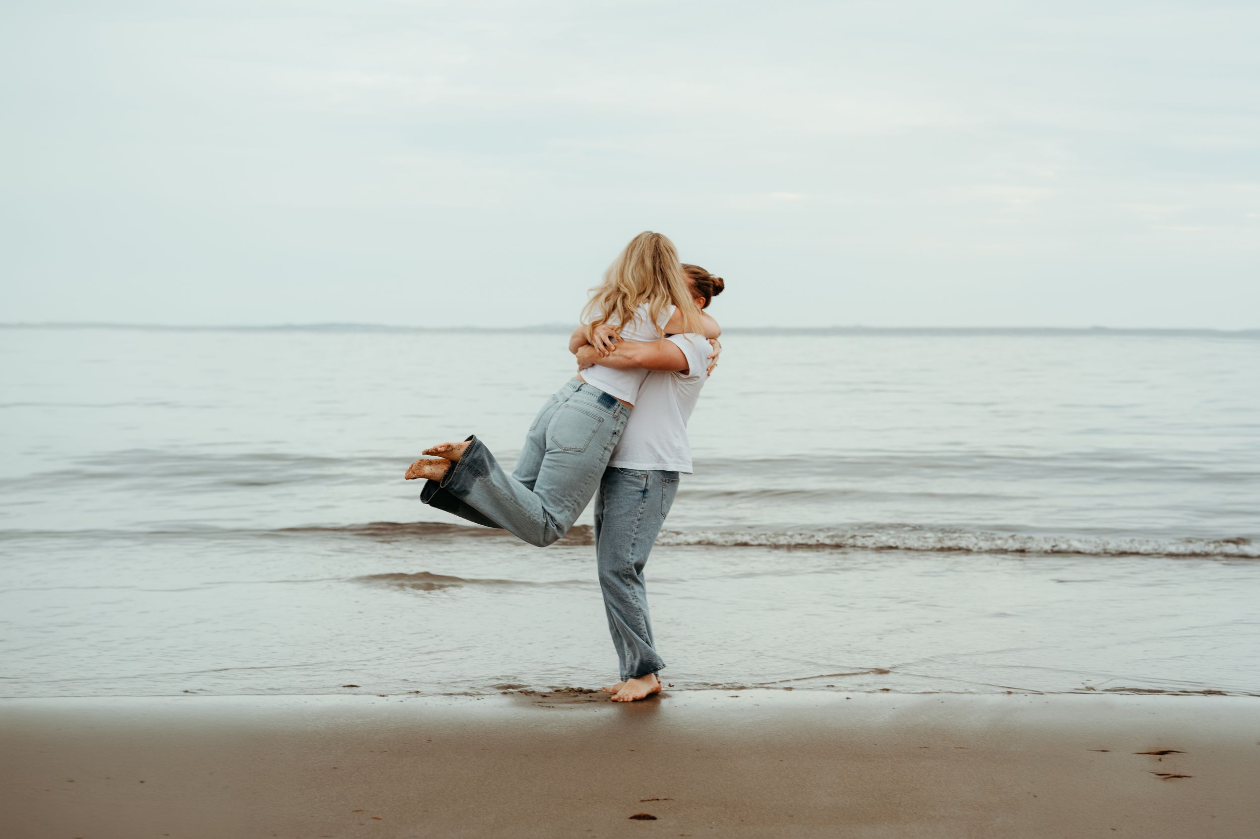 Couple embracing and spinning joyfully on a sandy beach with calm ocean waves in the background.