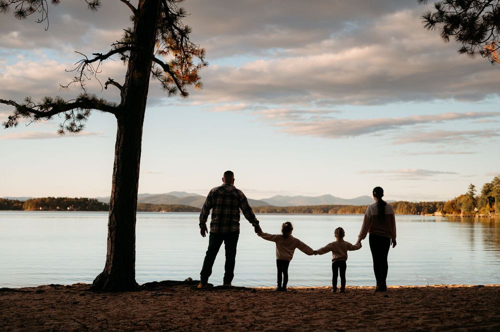 Family of four holding hands silhouetted against a serene lake at sunset.