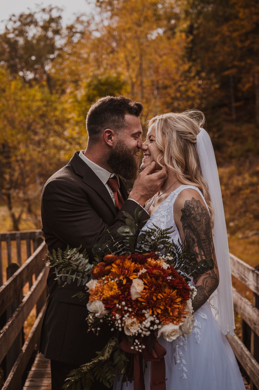 Bride and groom share a romantic moment on a wooden bridge surrounded by autumn foliage, holding a bouquet of fall flowers.