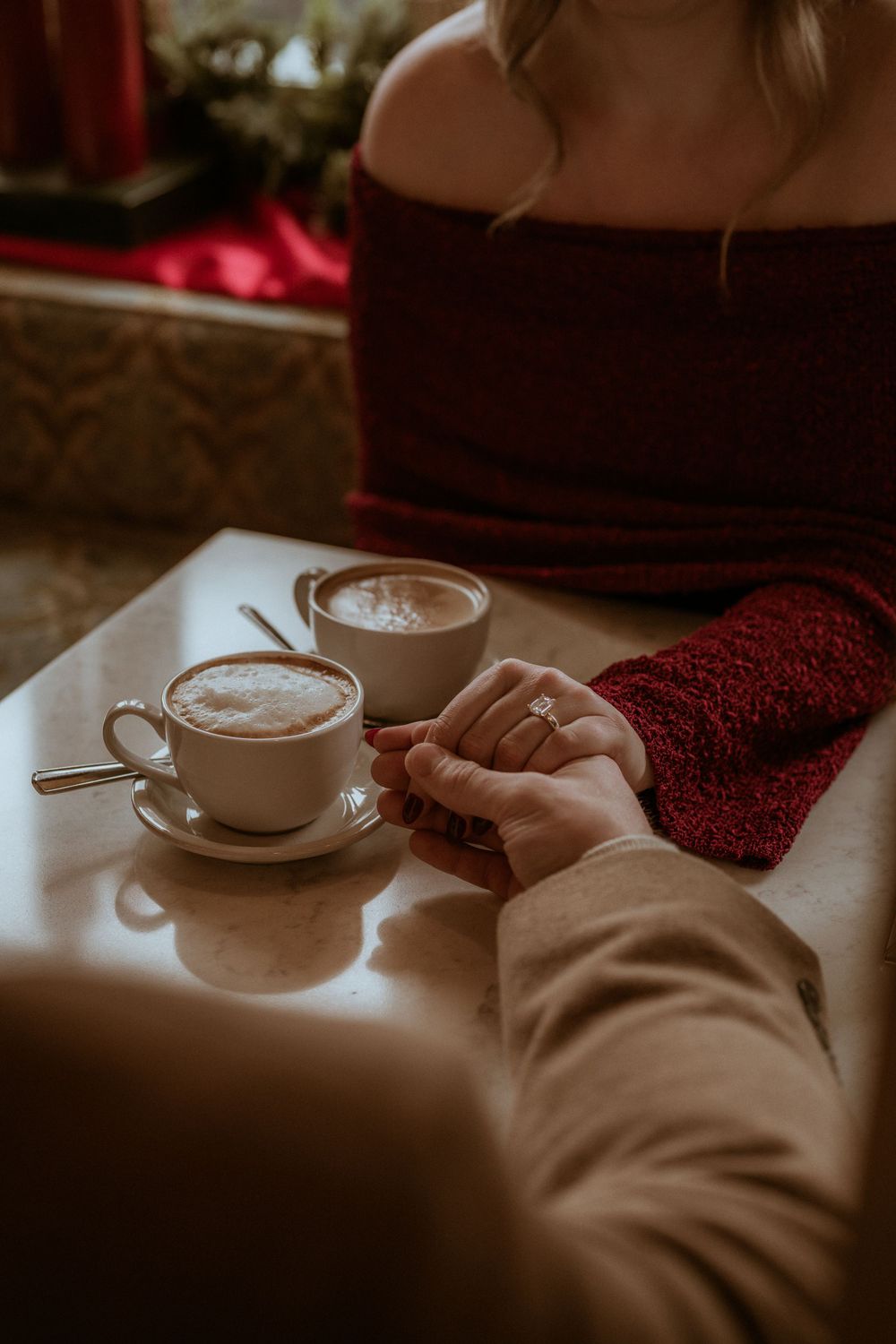 Couple holding hands over coffee cups at a cafe table with engagement ring visible.