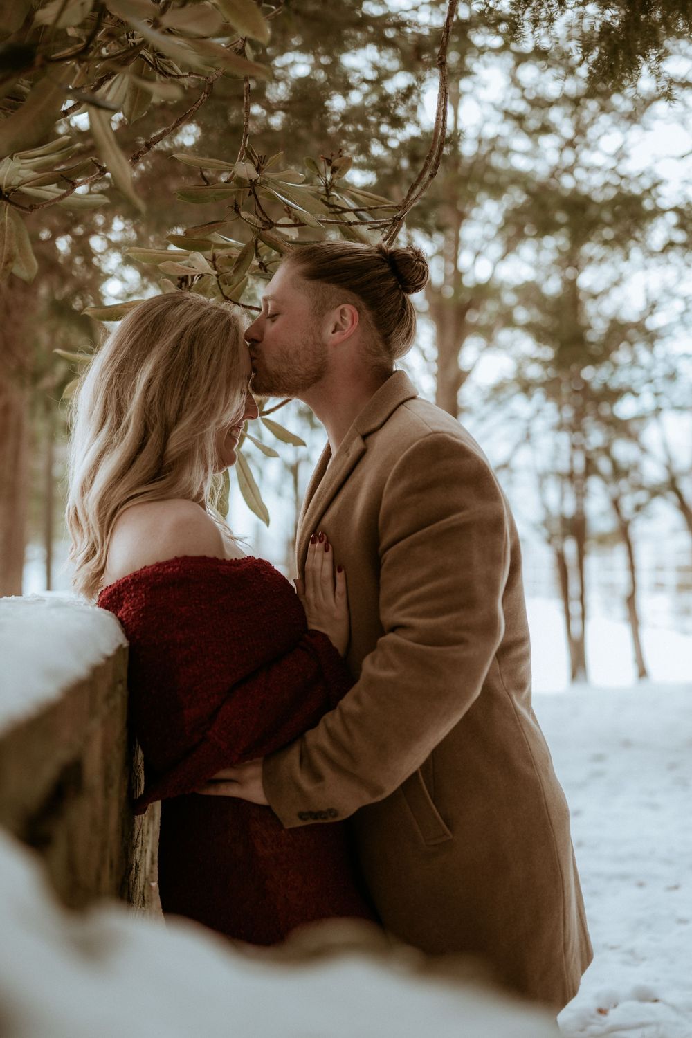 Couple kissing in a snowy forest, woman in red dress and man in tan coat.