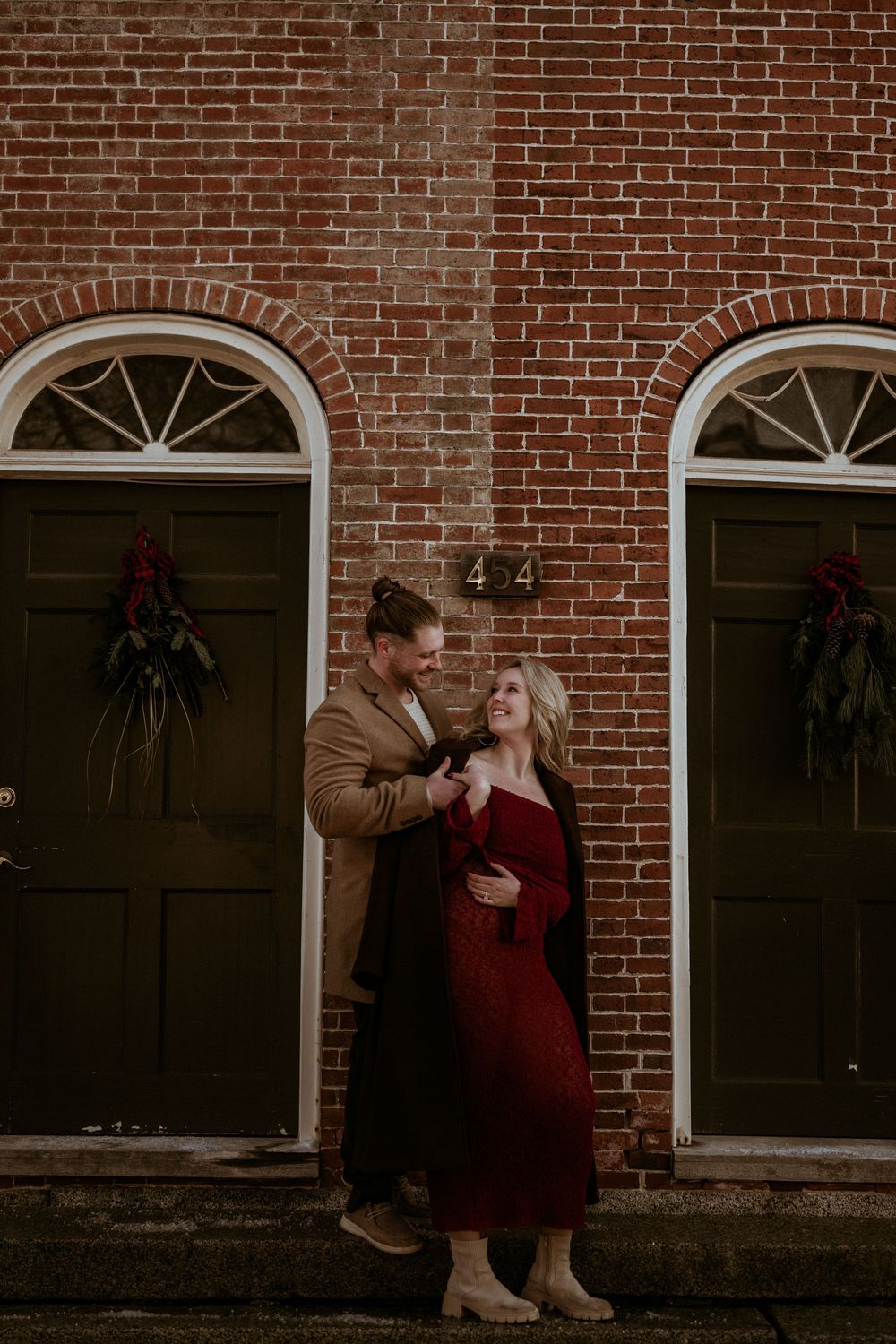 Couple embracing in front of red brick building with dark green doors adorned with holiday wreaths, woman in red dress.