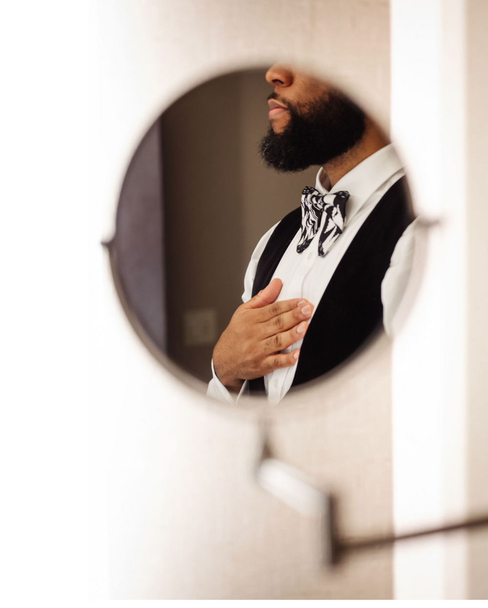 Groom adjusts his patterned bow tie while reflected in a round mirror before the wedding.