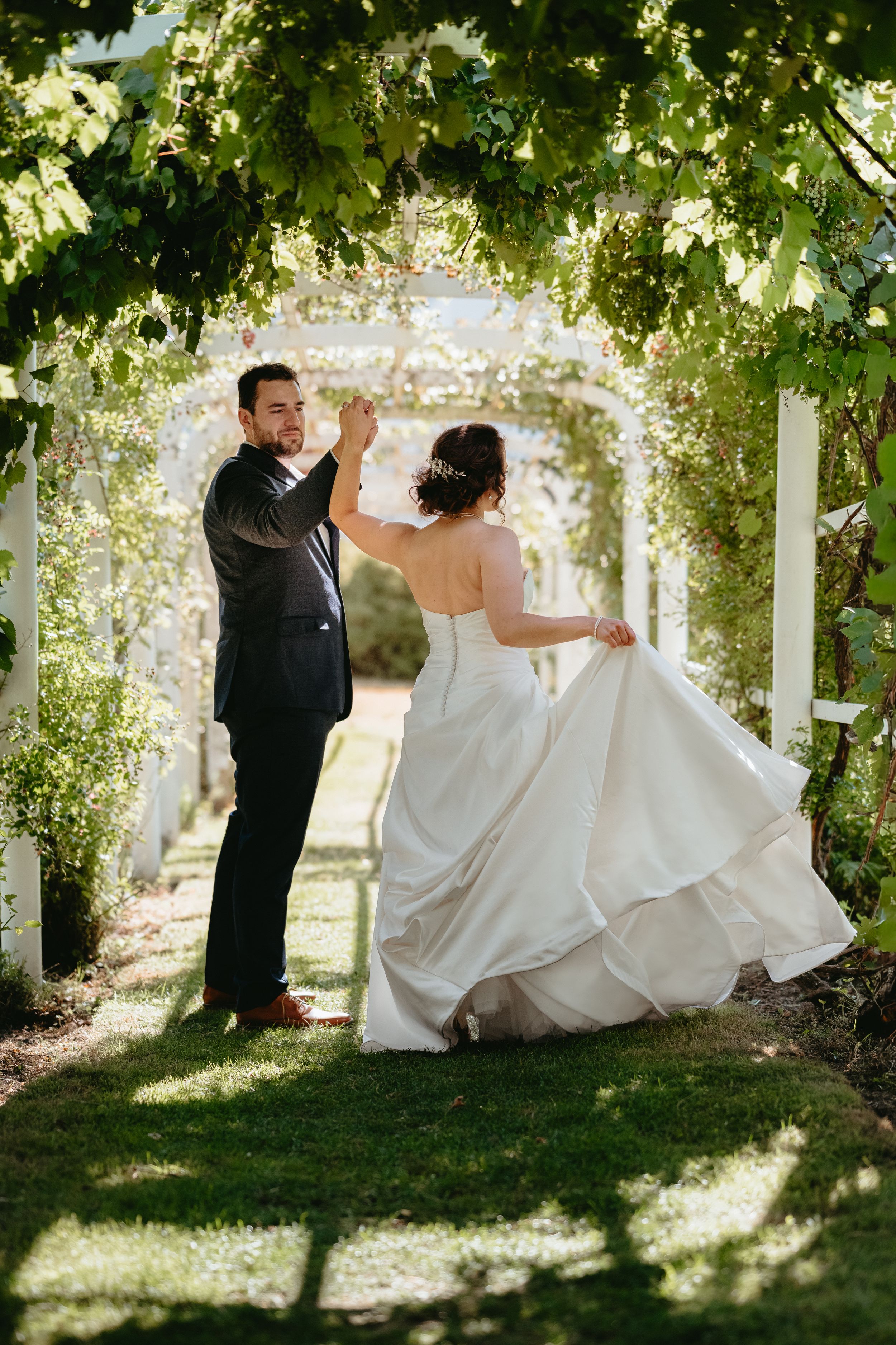 A bride and groom dance under a lush green pergola arch, the bride twirling in her elegant white gown.