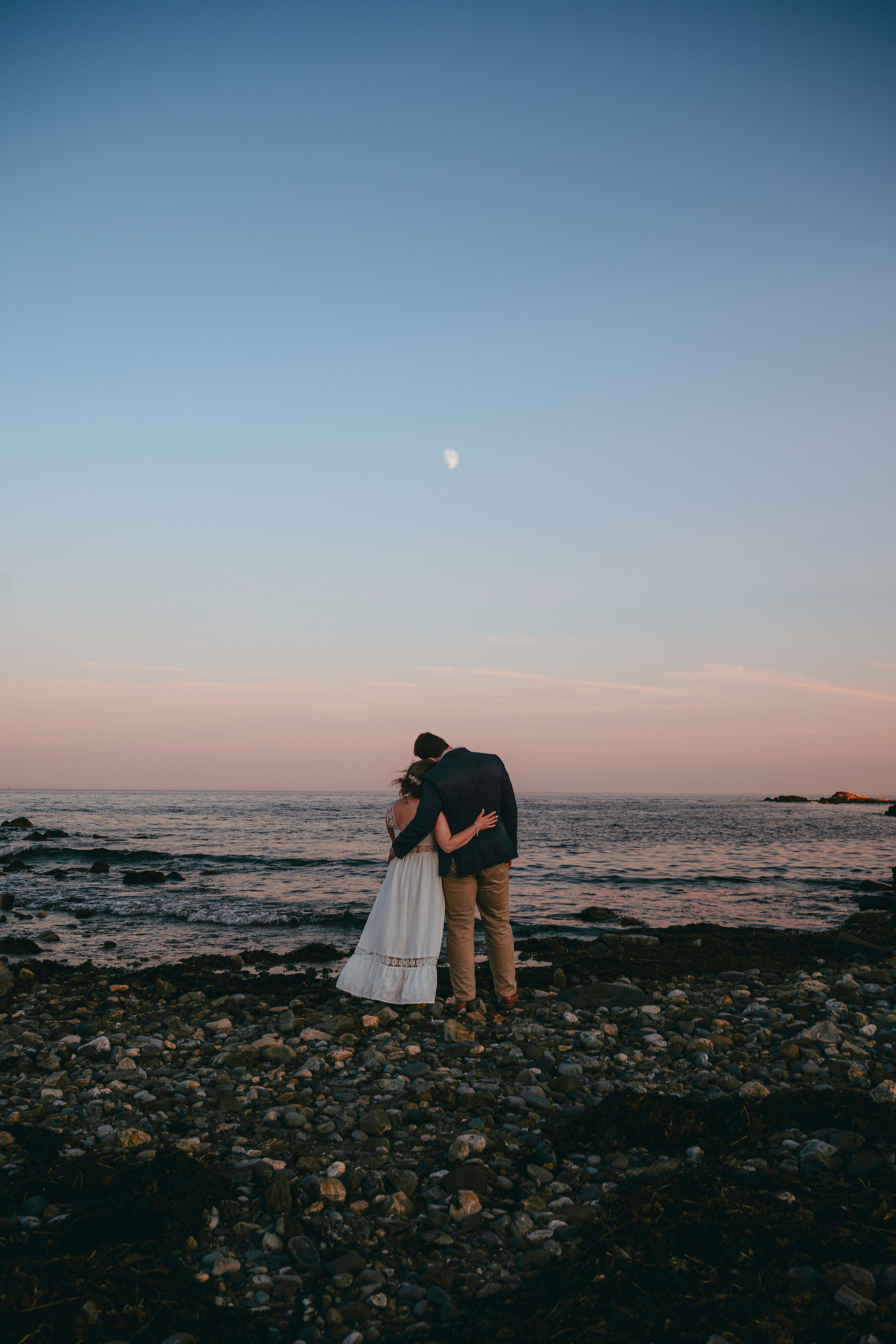 Couple embracing on rocky beach at sunset with moon visible in pastel sky over calm ocean waters.