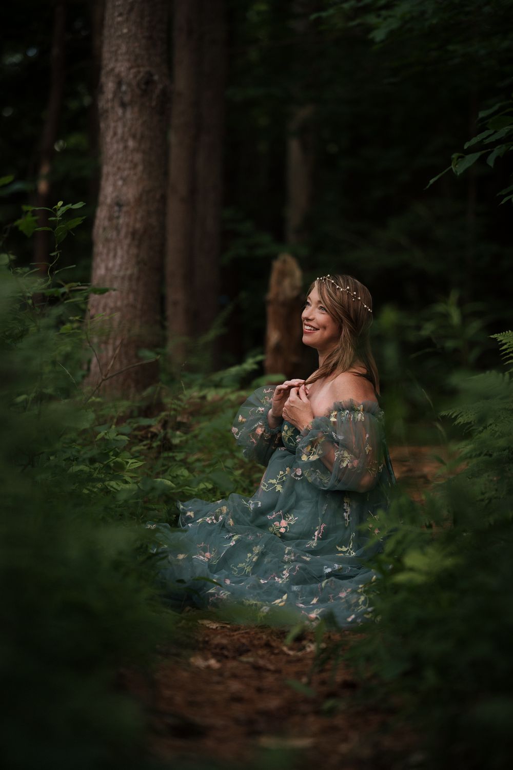 Smiling woman in teal floral off-shoulder gown with floral crown poses in lush green forest setting.