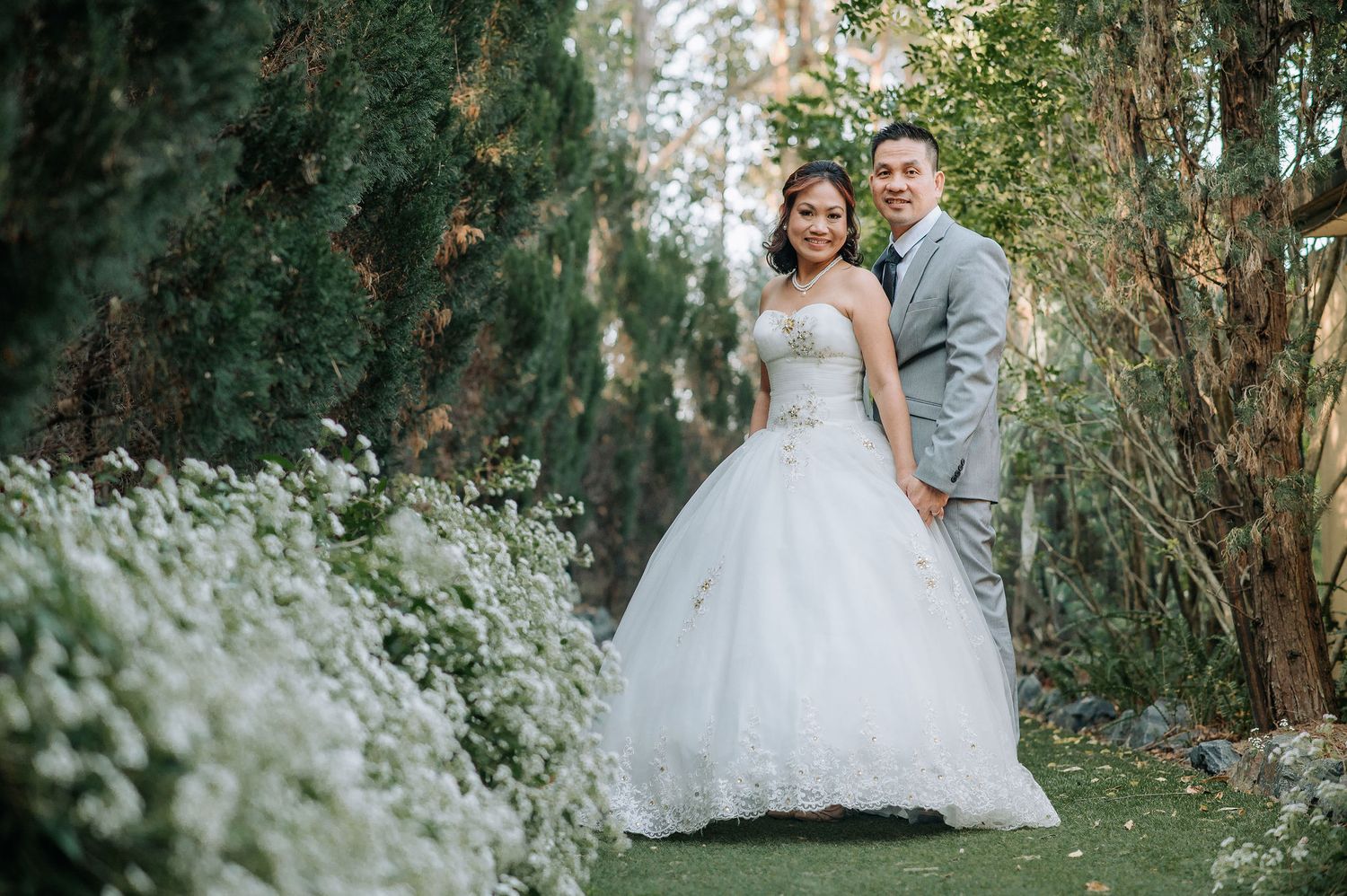 Wedding couple poses among white flowering bushes and trees in an intimate garden setting.