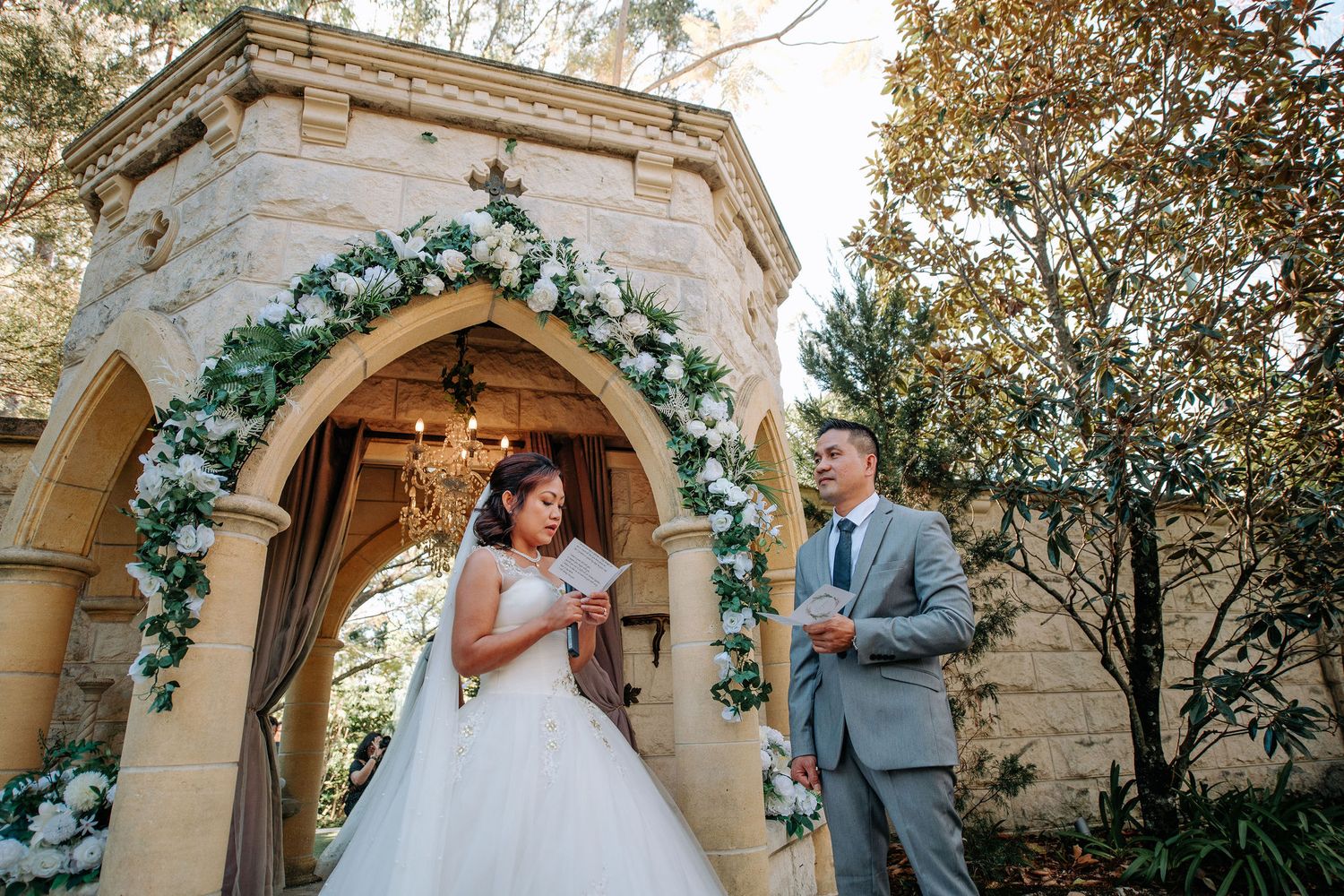 Wedding couple shares intimate moment under stone archway decorated with flowers.