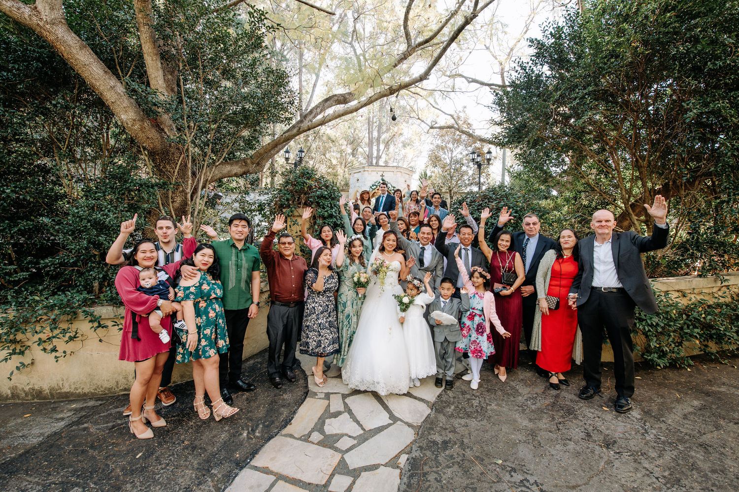 A festive wedding party group photo with guests wearing colorful attire on a stone path surrounded by greenery.