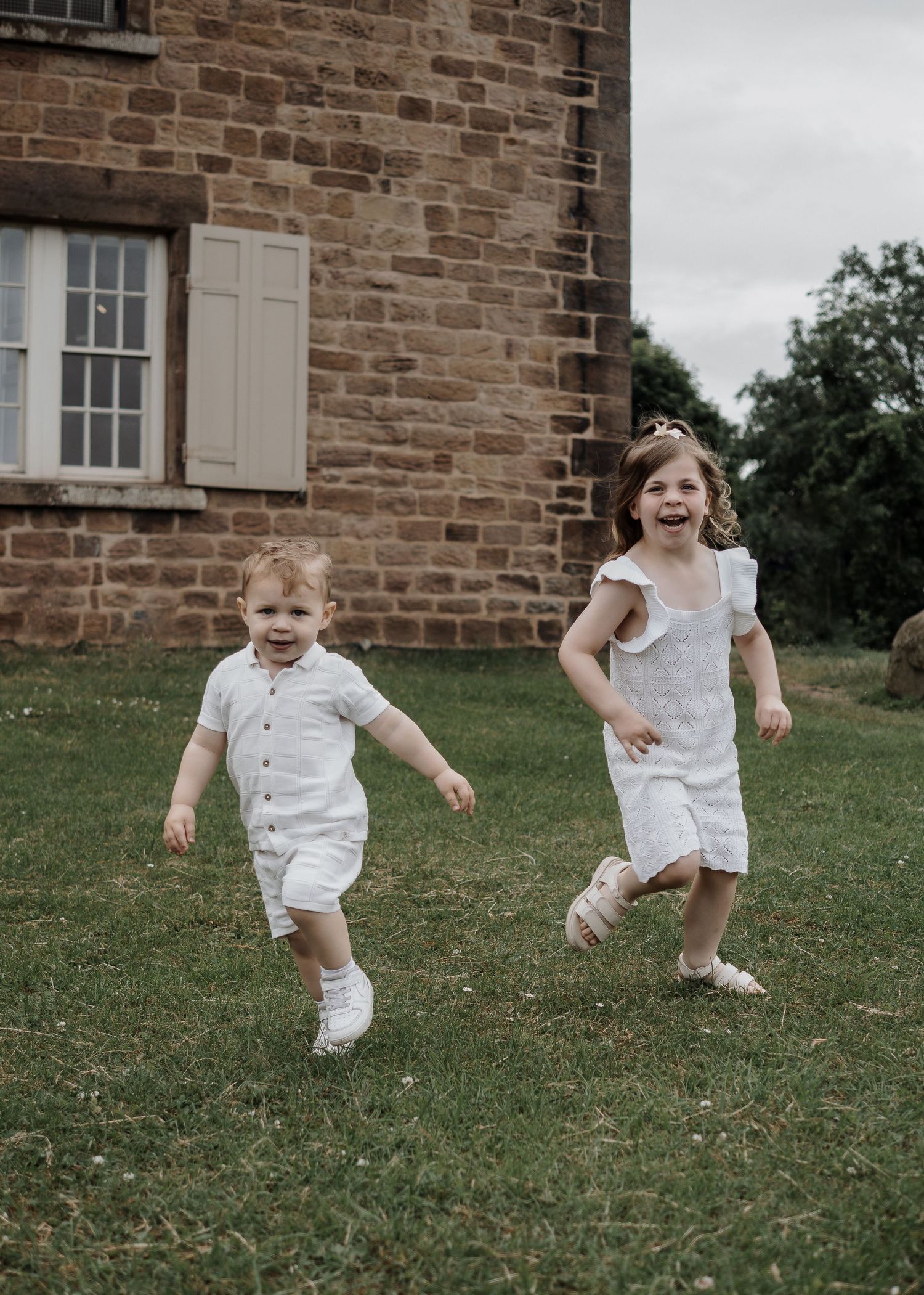 Family Photoshoot - Children in white outfits jump and play on a grassy lawn near a brick building.