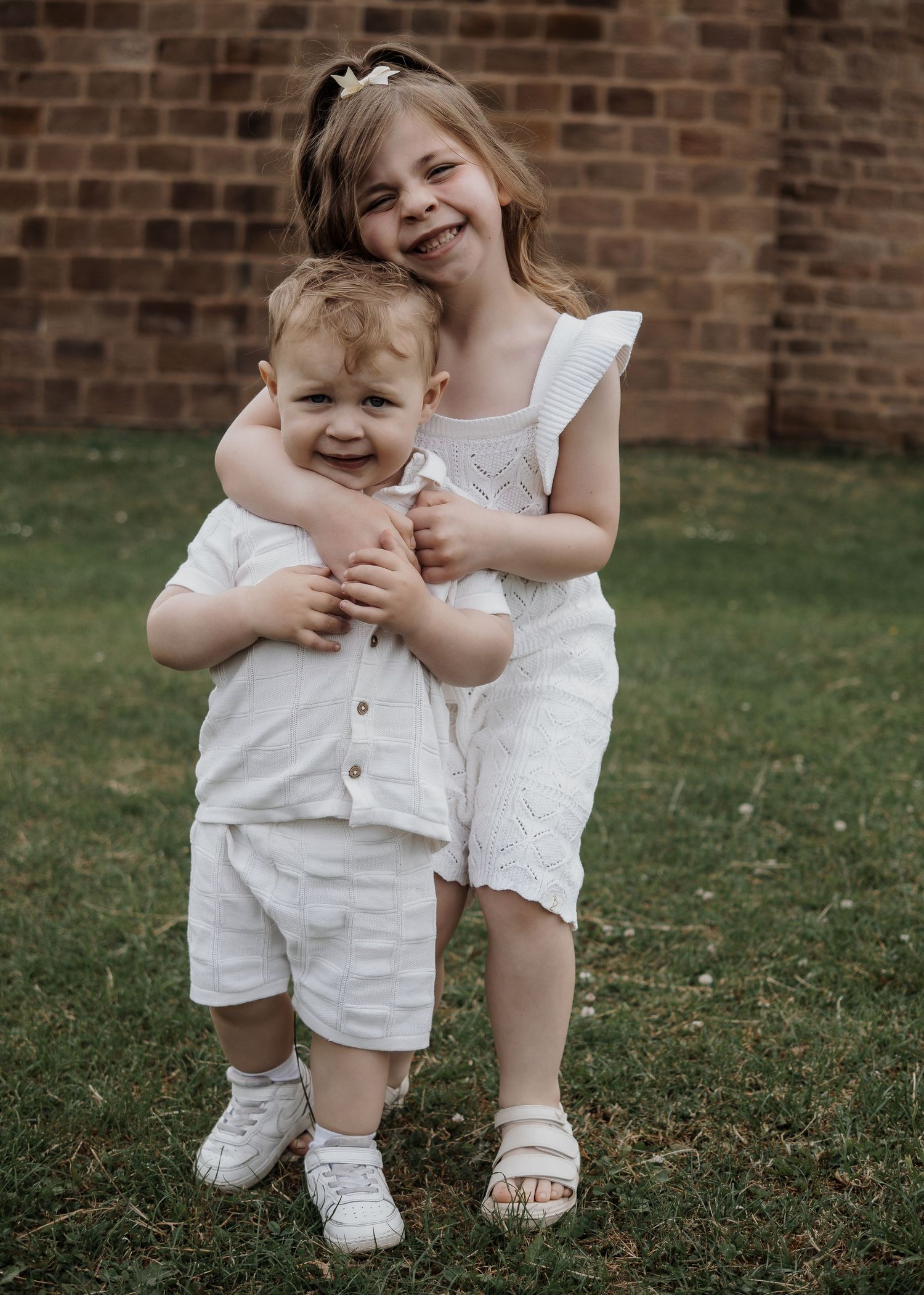 Family Photoshoot - Two people in white summer outfits share a tender moment on grass near a brick wall.