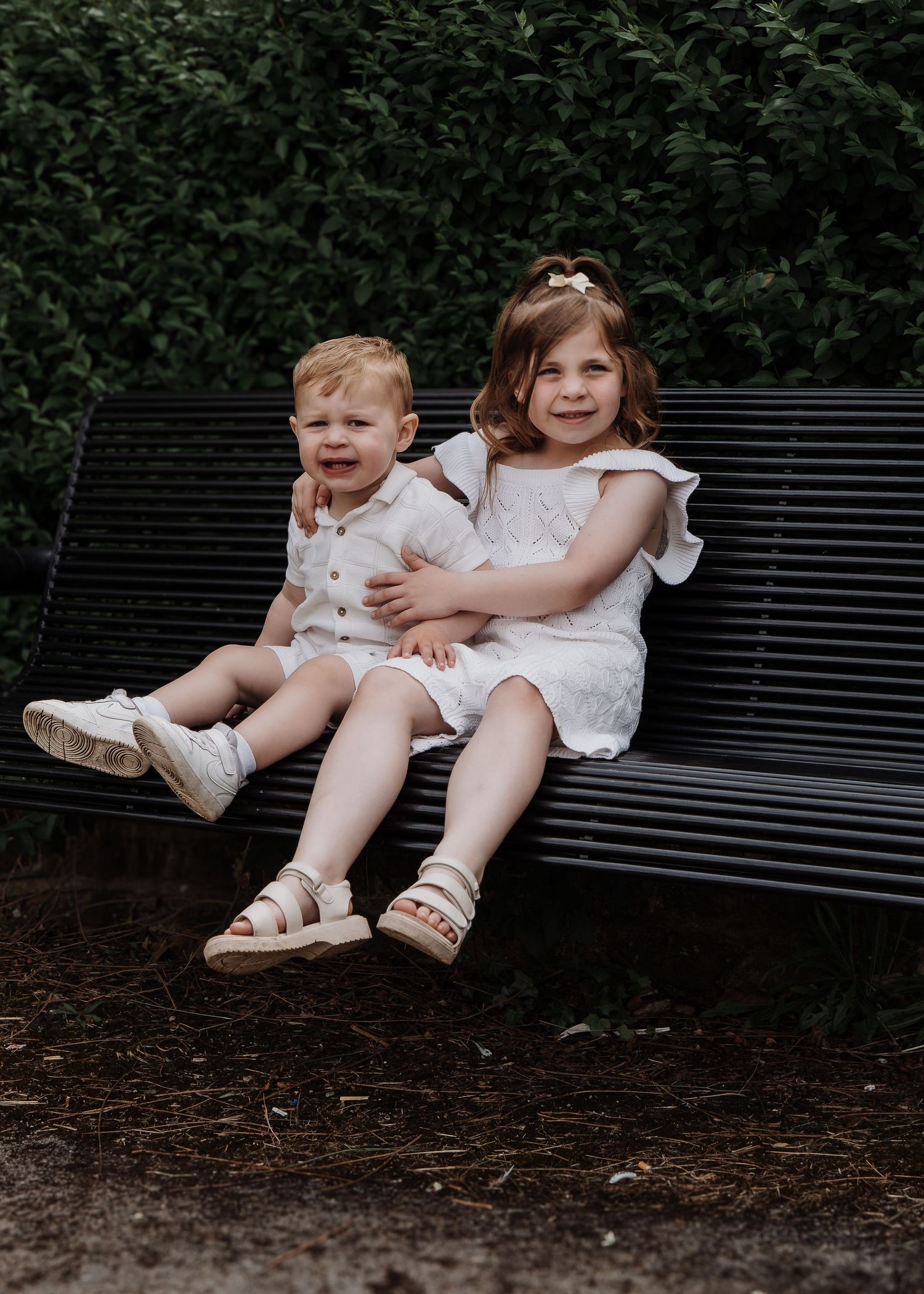 Family Photoshoot - Two people in white outfits sit together on a black park bench surrounded by greenery.