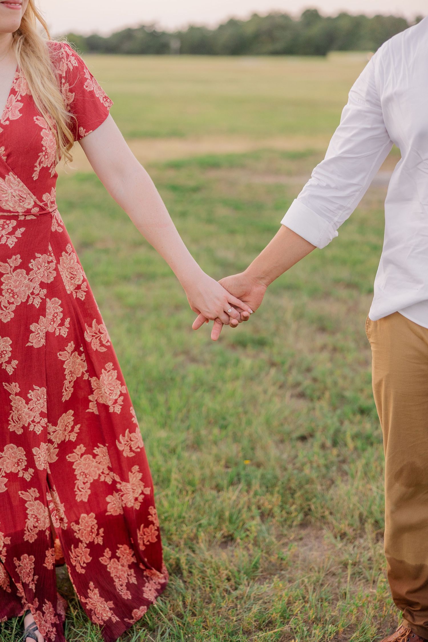Couple holding hands in field wearing red floral dress and white shirt.