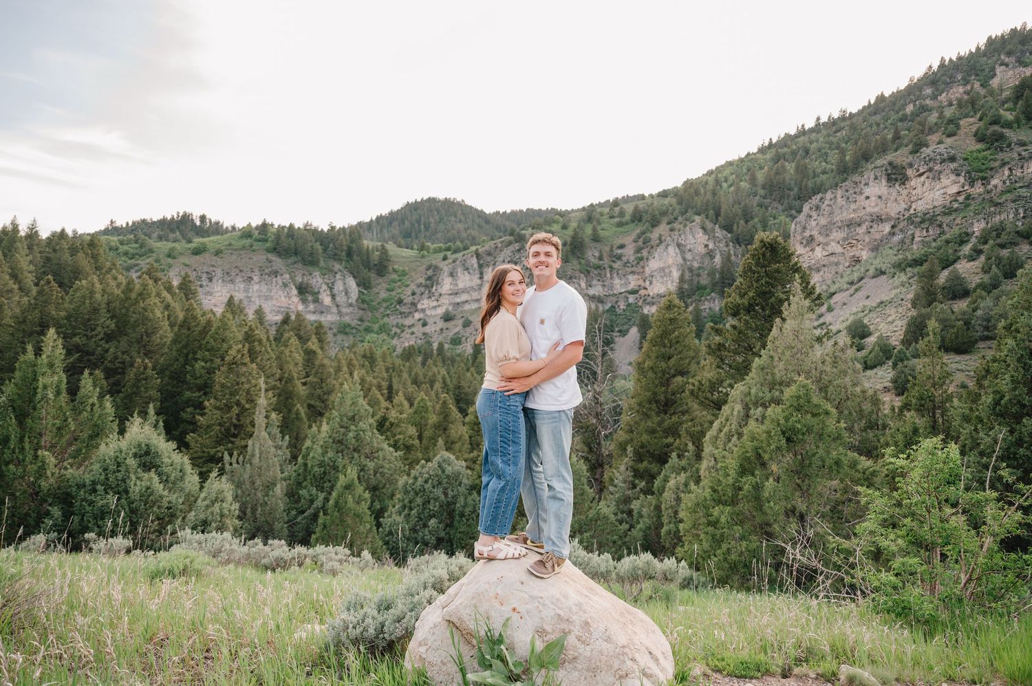 A couple stands together on a large boulder against a backdrop of forested mountains and cliffs.