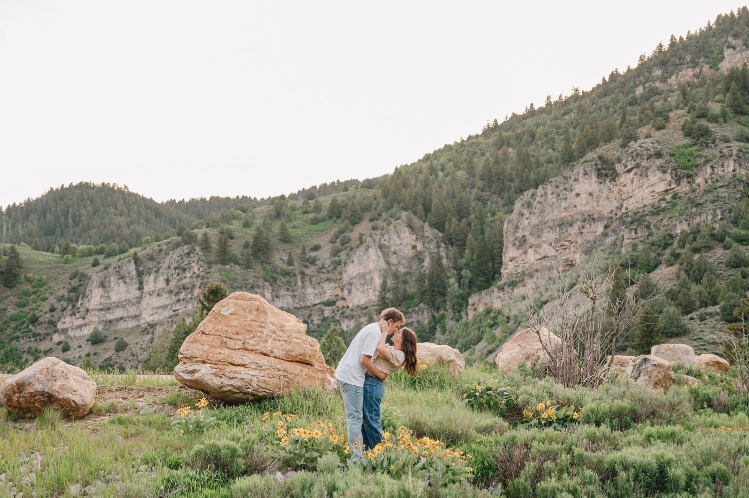 A couple embraces while standing among rocky outcrops and green vegetation against towering mountain cliffs.