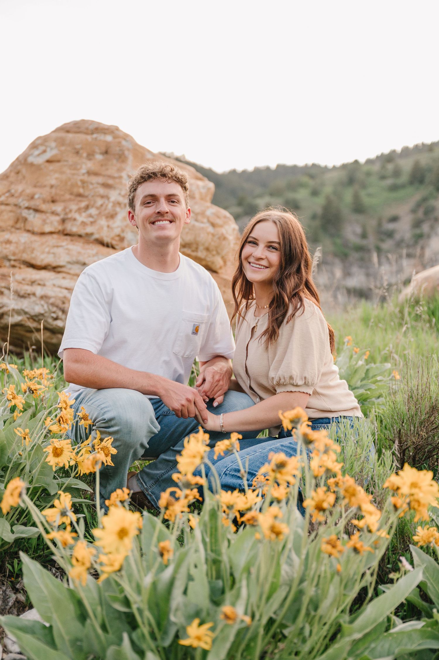 Couple sits in scenic mountain landscape surrounded by blooming yellow daylilies and red rock formations.