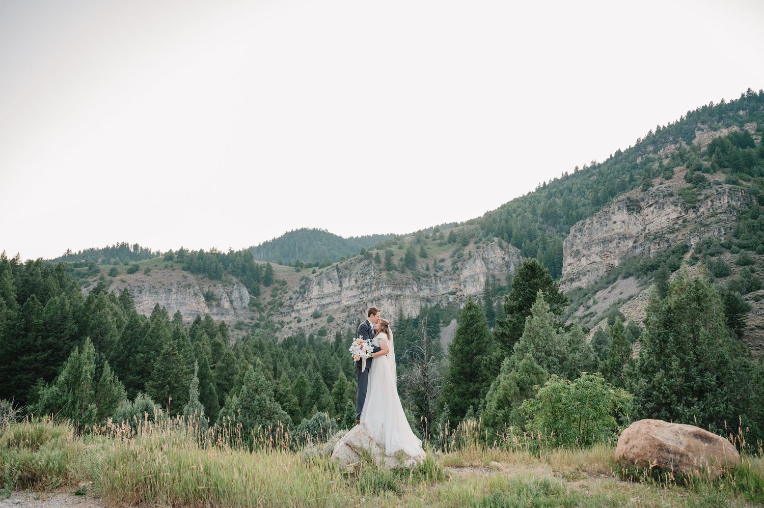 A wedding couple poses in a scenic mountain location with dramatic cliff faces in the background.