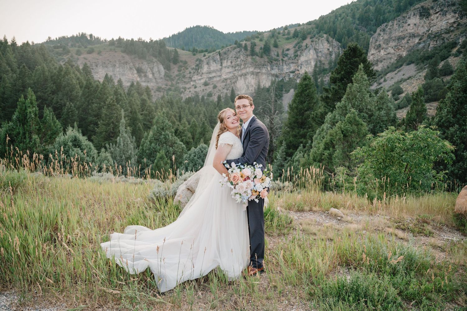 A bride and groom embrace in a mountain meadow with rocky cliffs behind them.