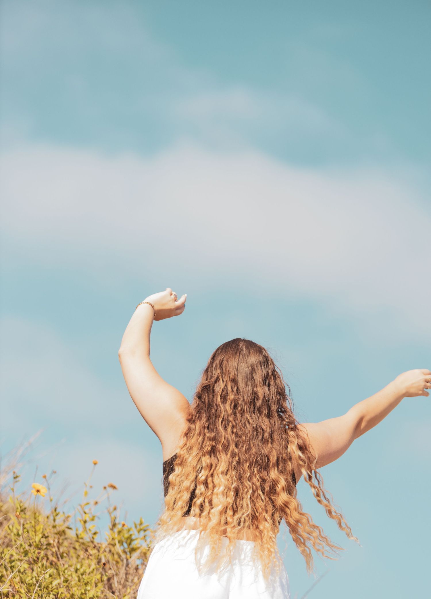 Person with long curly hair wearing white dress raises arms towards blue sky with yellow wildflowers in foreground.