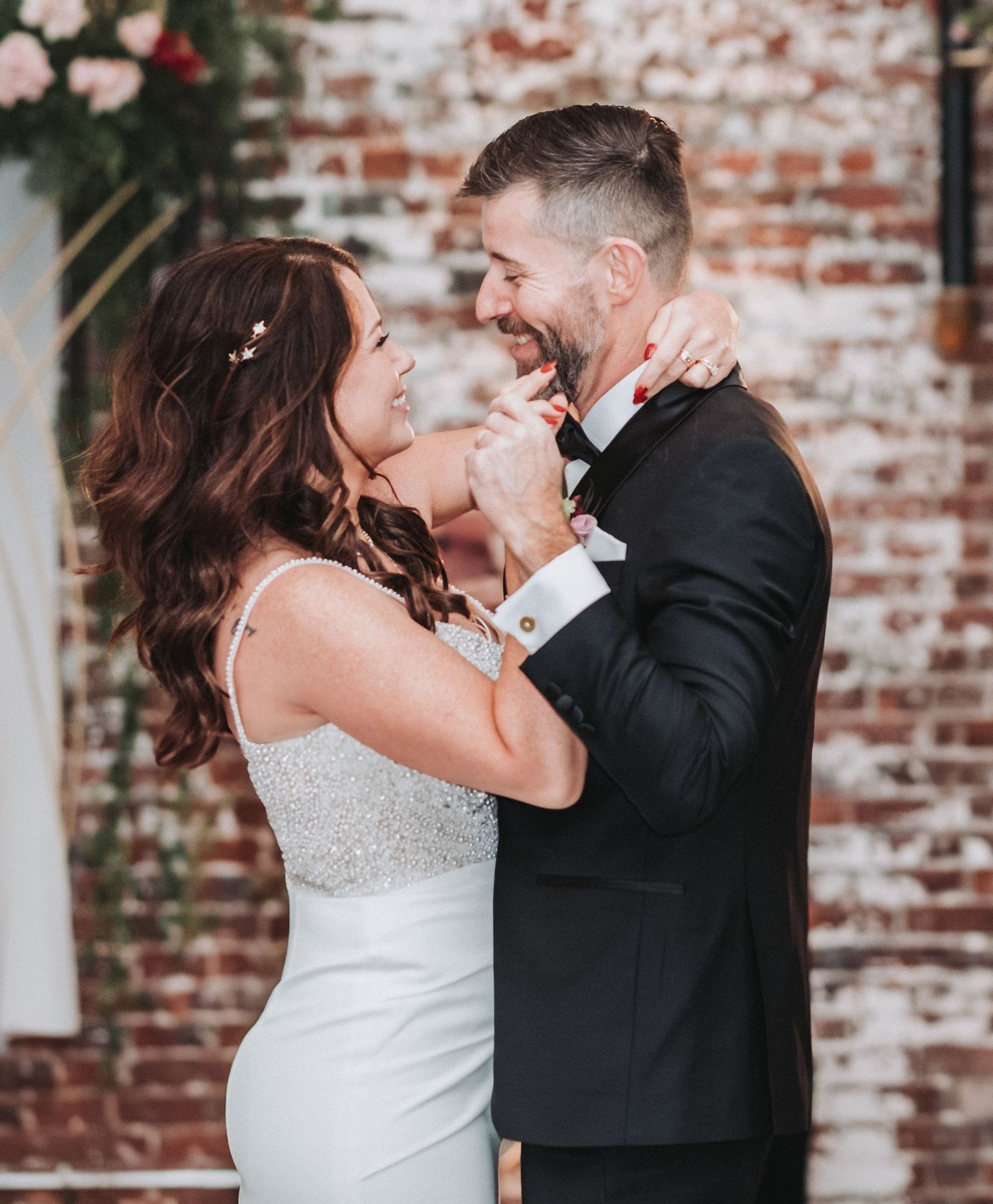 A joyful couple in wedding attire share an intimate moment while laughing against a rustic brick wall backdrop.