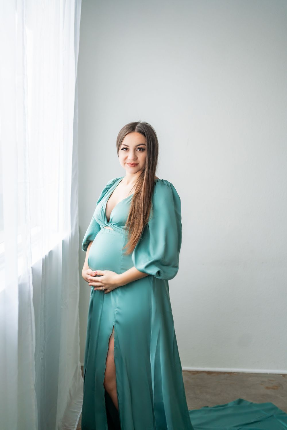 A maternity portrait featuring an elegant teal silk dress in a minimalist white room setting.