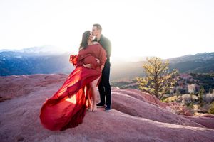 Couple embraces on red rocks during sunset with flowing red dress catching wind.