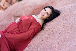 Woman poses on red rocks wearing flowing crimson dress during maternity photoshoot.