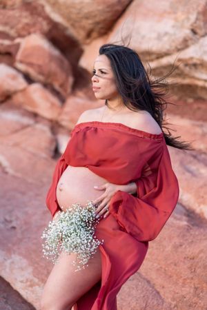 A maternity photoshoot in a red off-shoulder dress against red rocks while holding baby's breath flowers.