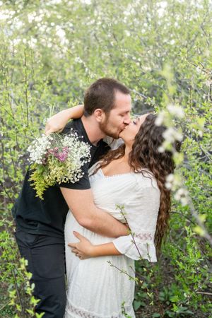 A romantic embrace between a couple surrounded by white flowering bushes in a natural setting.