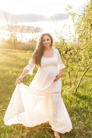 A woman in a flowing white dress twirls in a sunlit meadow with trees in the background.