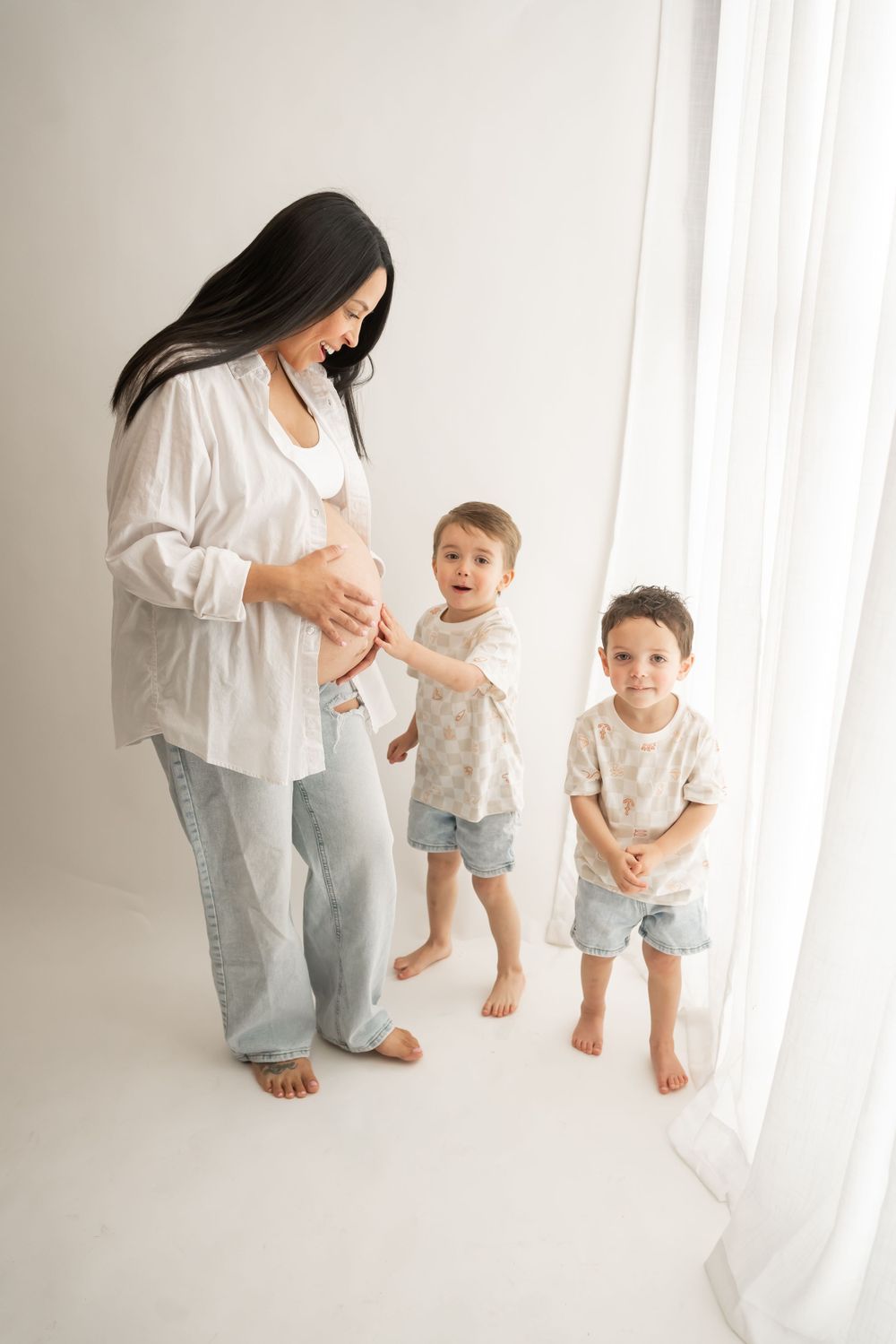 Pregnant woman in white shirt standing with two small children by window.