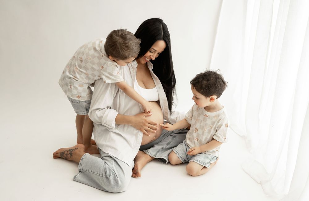 Pregnant woman in white shirt playing with two children on floor by window.