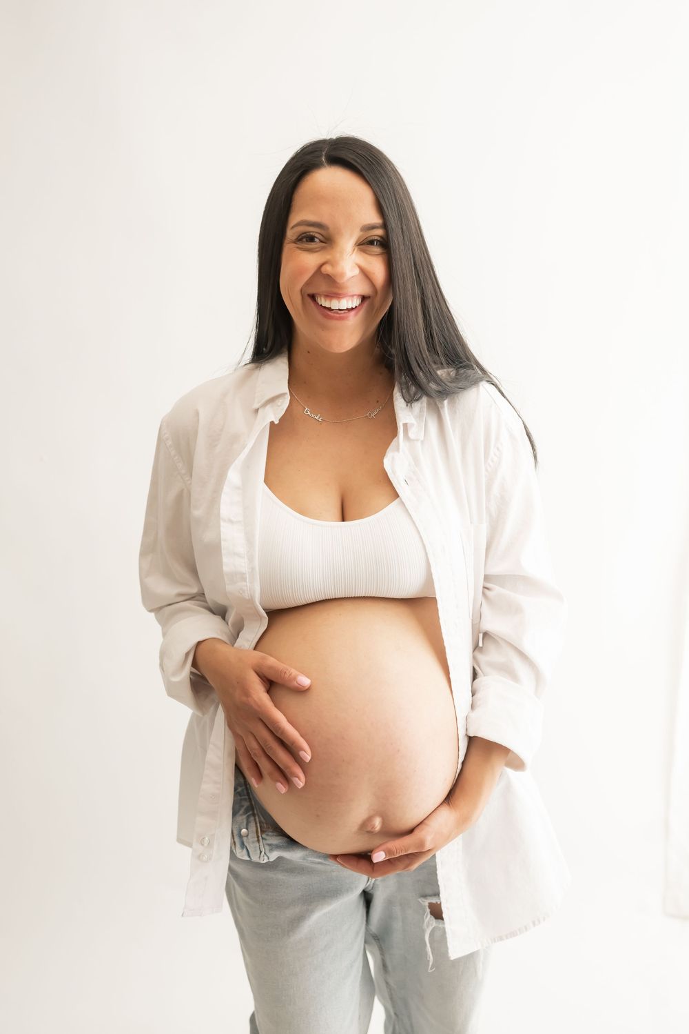 Pregnant woman in white button-down shirt and jeans smiling at camera.