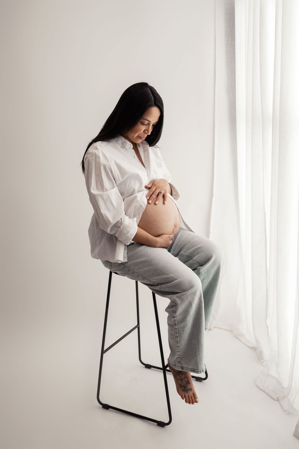 Expectant mother in white shirt and gray pants sitting on stool near window.