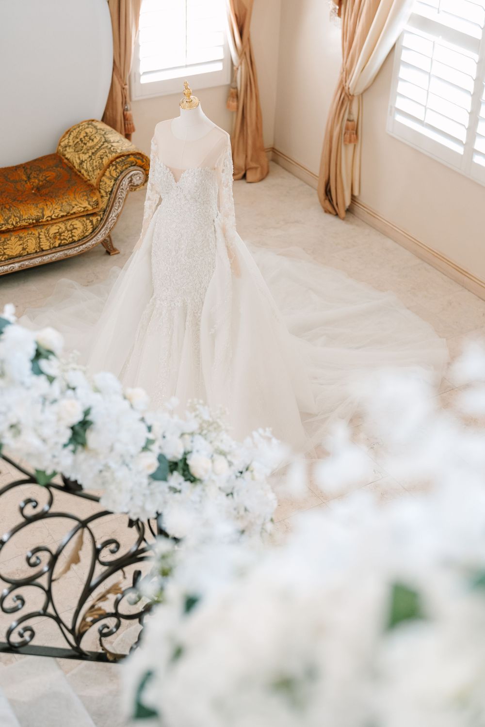 Elegant white wedding dress displayed in a sunlit room with delicate white floral decorations and beige curtains.