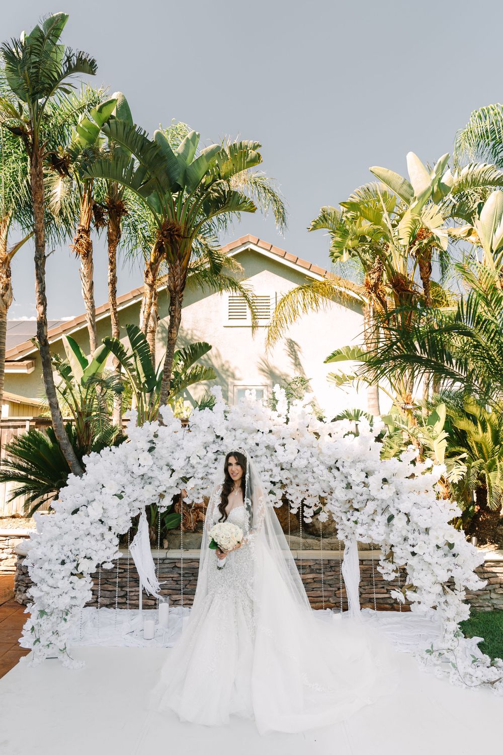 White floral wedding arch decorated with orchids stands elegantly against a backdrop of tropical palm trees.