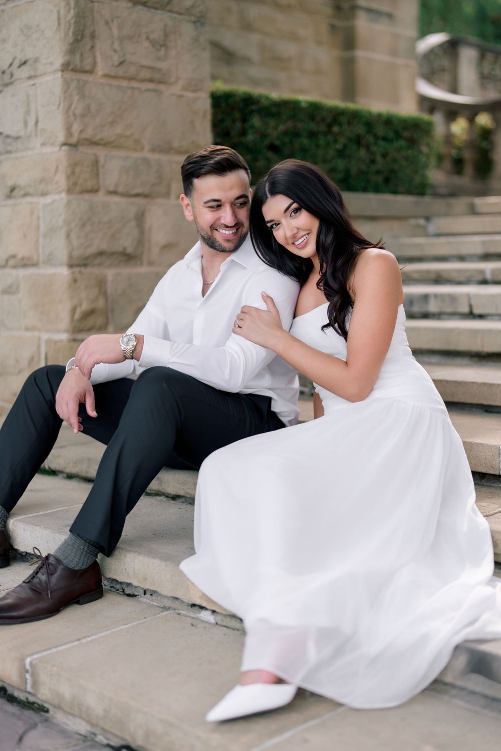 A smiling couple in white outfits sits on stone steps, with the woman leaning against the man in an elegant outdoor setting.