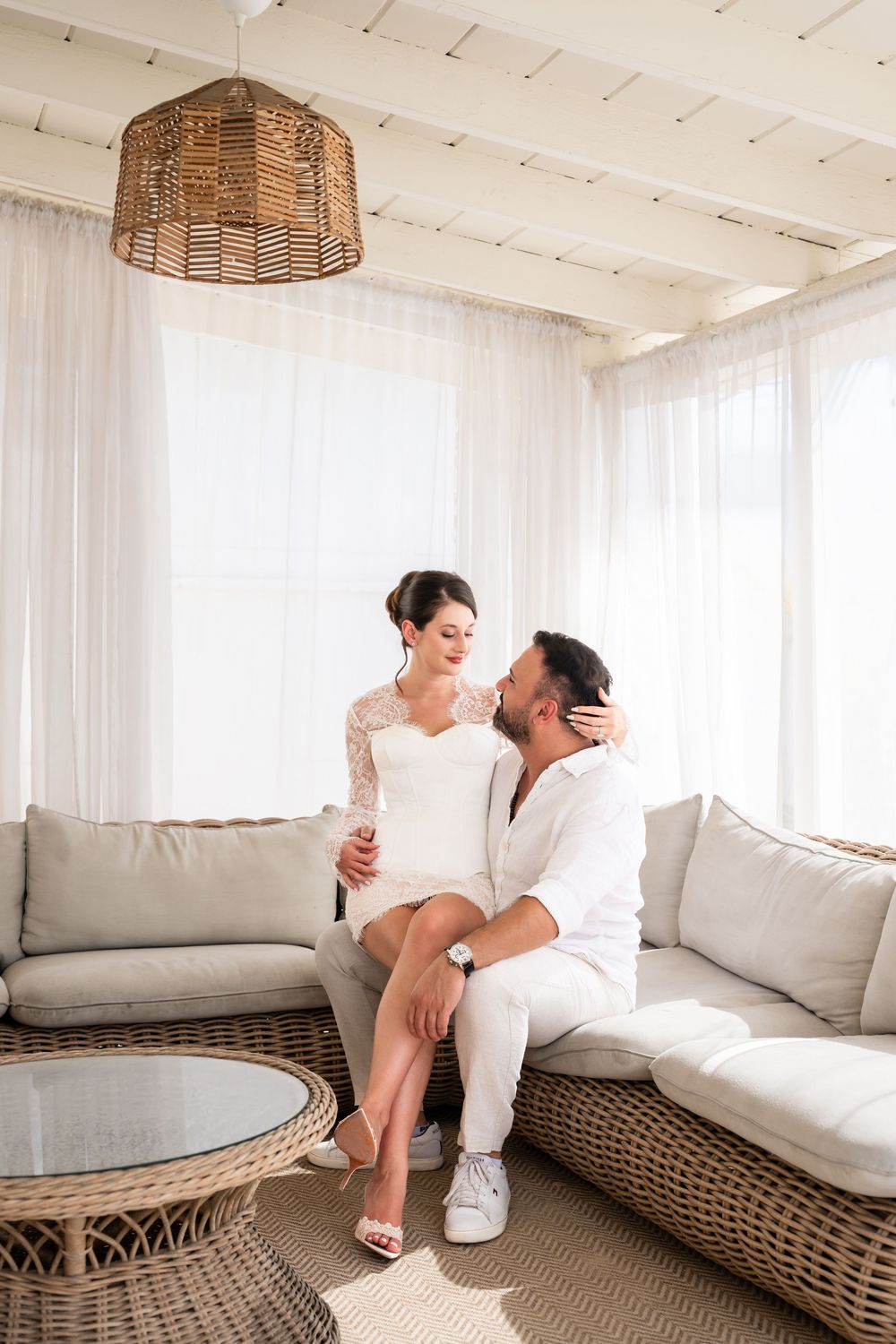 Bride and groom sitting together on a wicker sofa in a bright, airy room with white curtains and natural light.