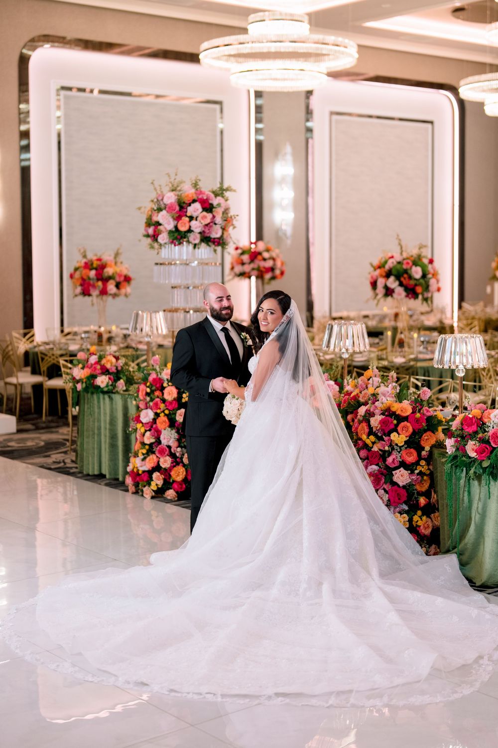Bride in flowing white gown with veil and groom in black suit pose in elegant floral wedding reception venue.