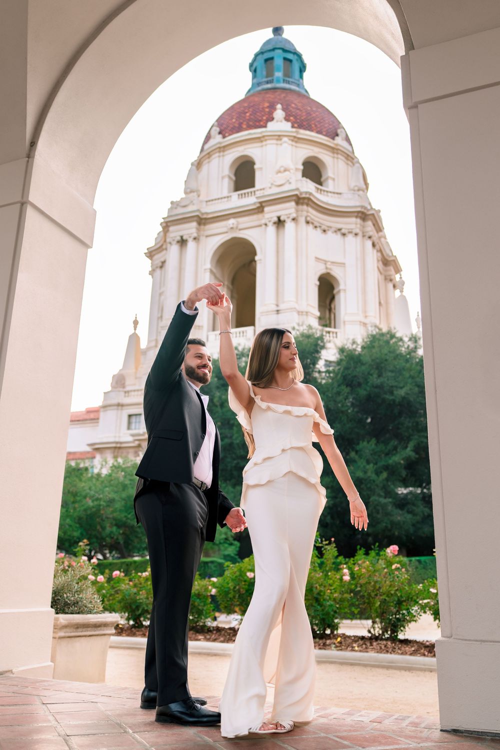 A couple in formal attire dances and poses beneath white archways at a historic church courtyard with domed towers.