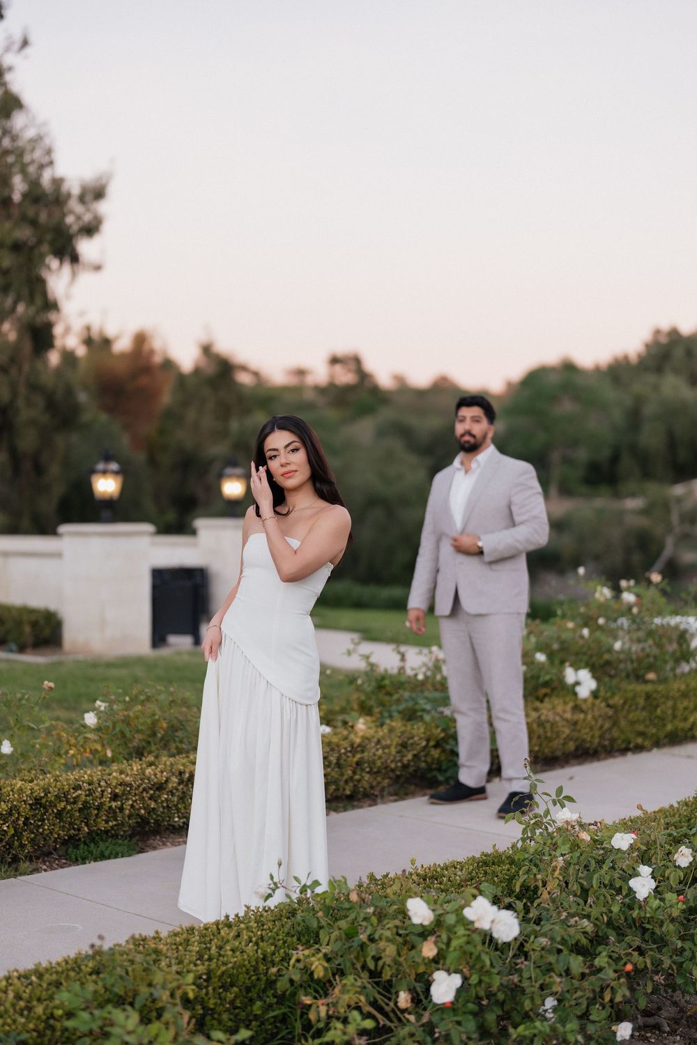 A couple in elegant white attire pose in a garden with blooming white roses at sunset.