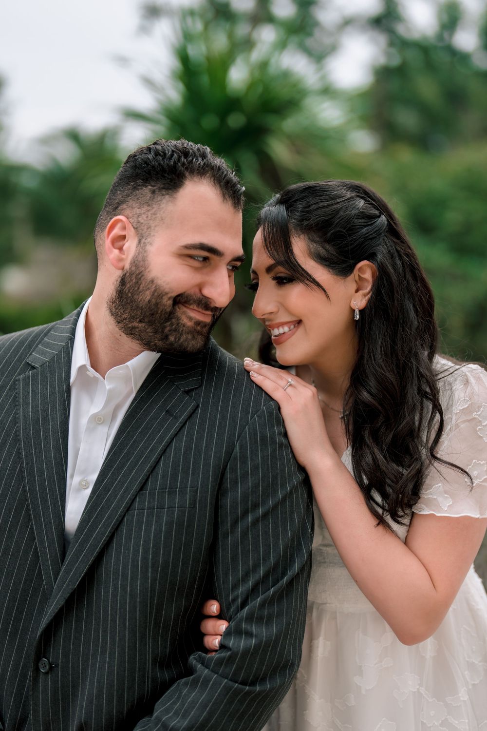 A romantic couple shares an intimate moment outdoors, with the woman in white and man in pinstripe suit smiling together.