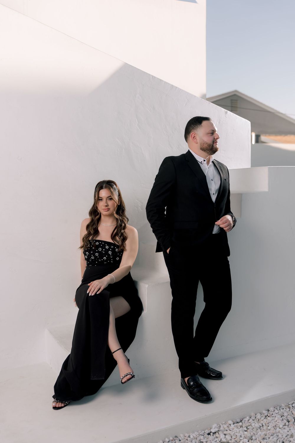 A couple in black formal attire poses artistically on white steps for a modern minimalist photoshoot.