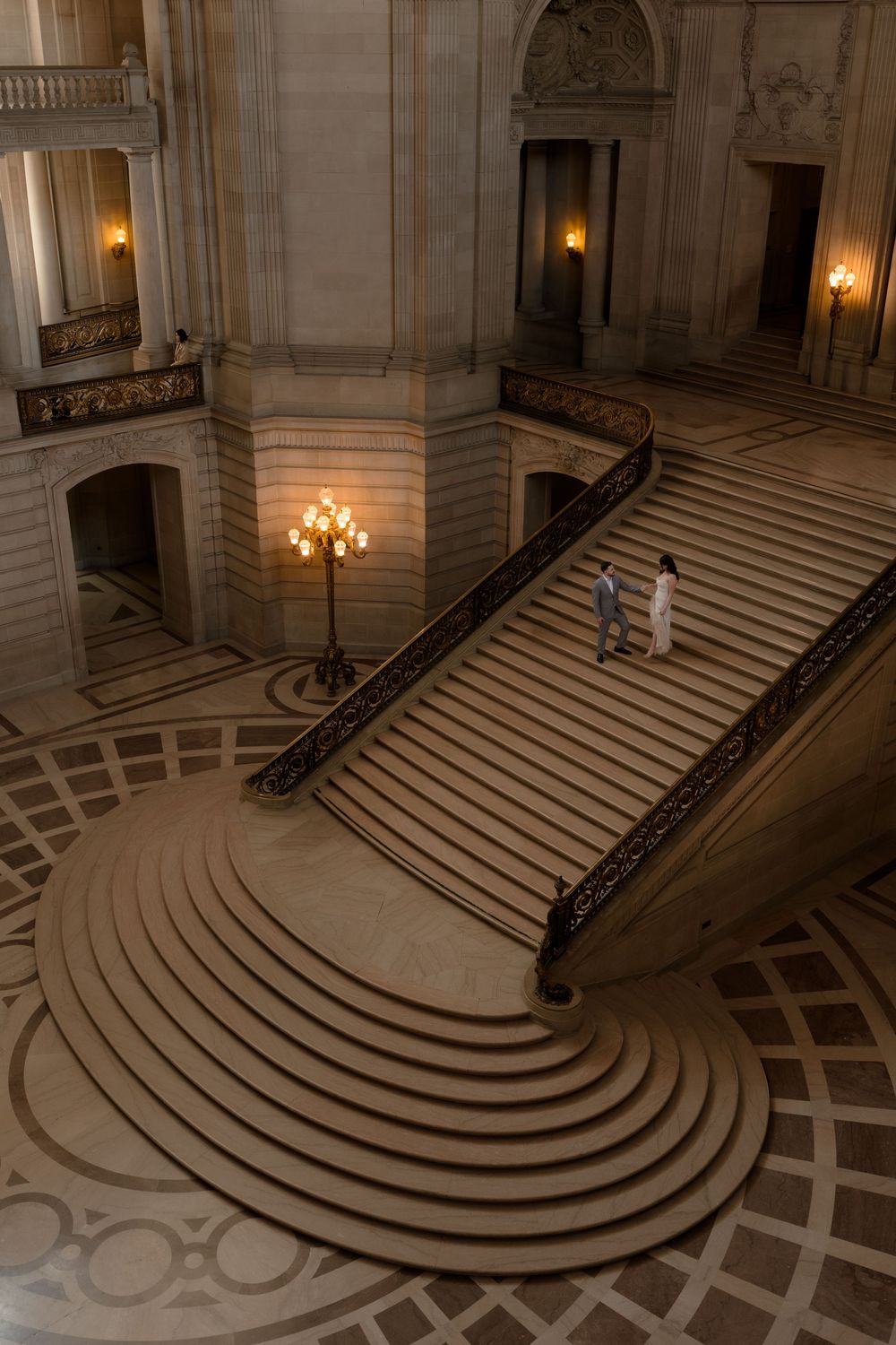 Couple standing on grand curved staircase inside San Francisco City Hall, captured from above with warm golden lighting.