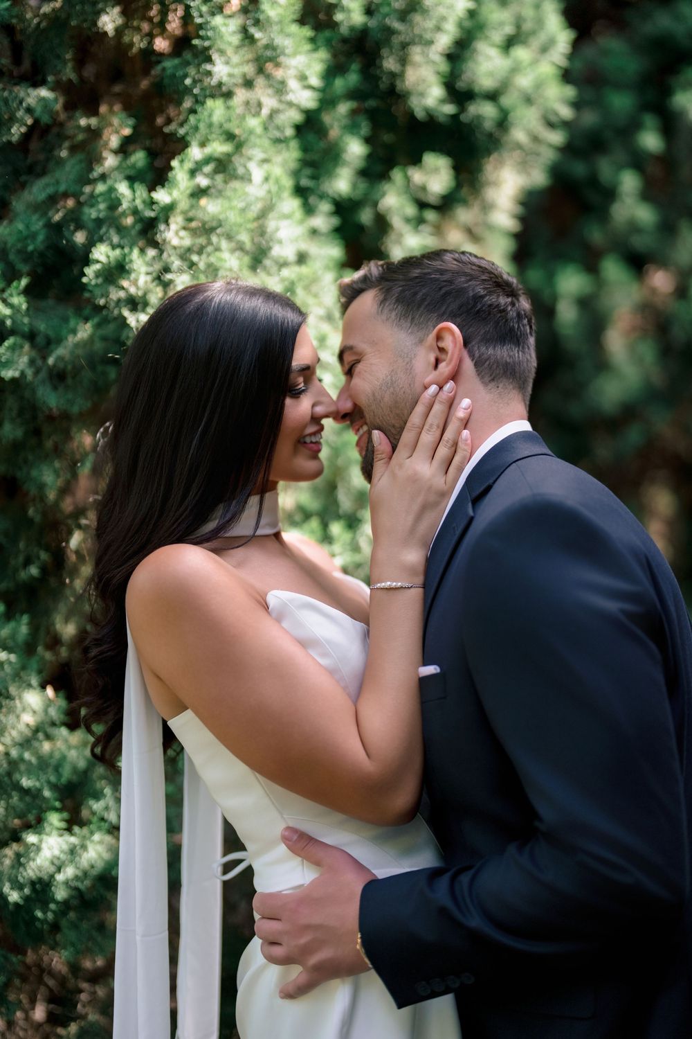 Bride and groom sharing a romantic moment near white columns surrounded by green foliage.